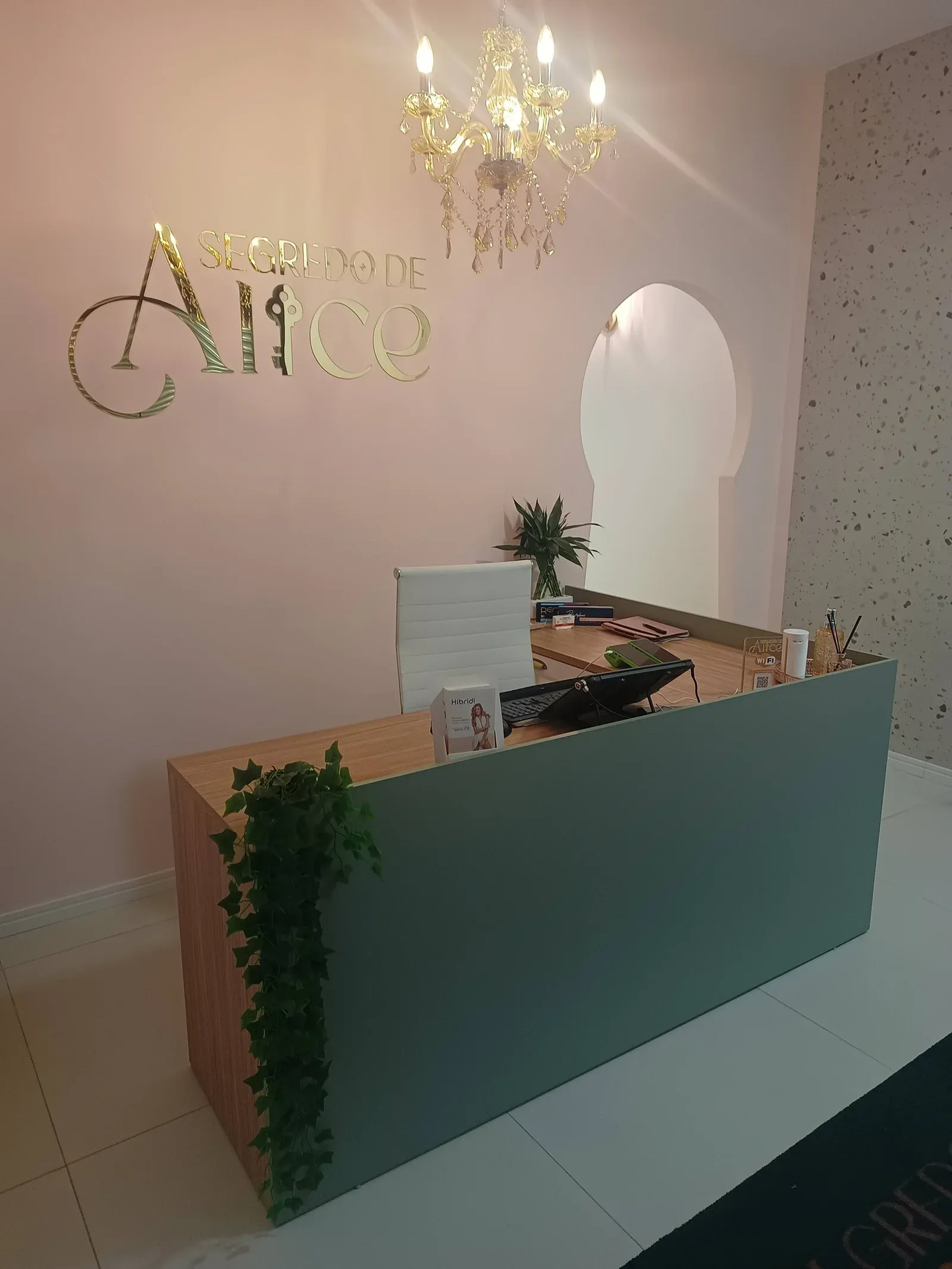 Recepção da Segredo de Alice: parede rosa com o logo dourado, lustre de cristal e balcão verde